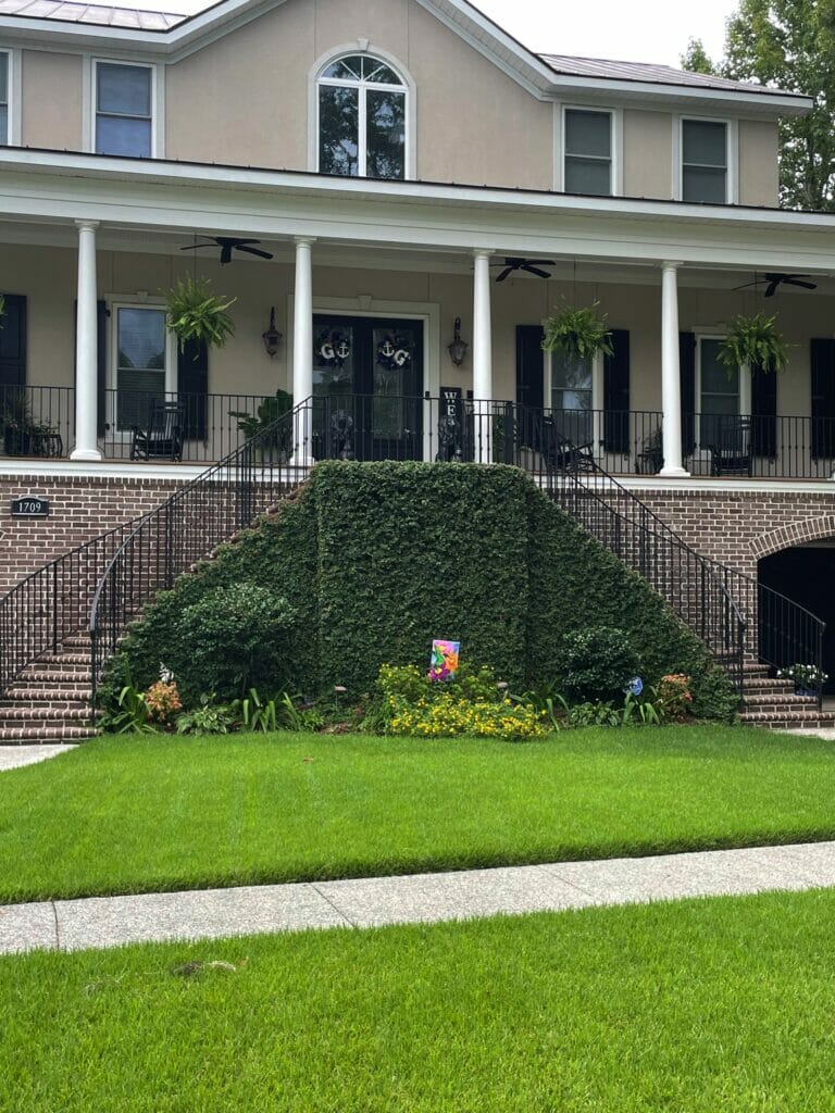 Two-story house with large front steps covered in green ivy, symmetrical porch columns featuring custom wrought iron work, hanging plants, and a well-manicured lawn.