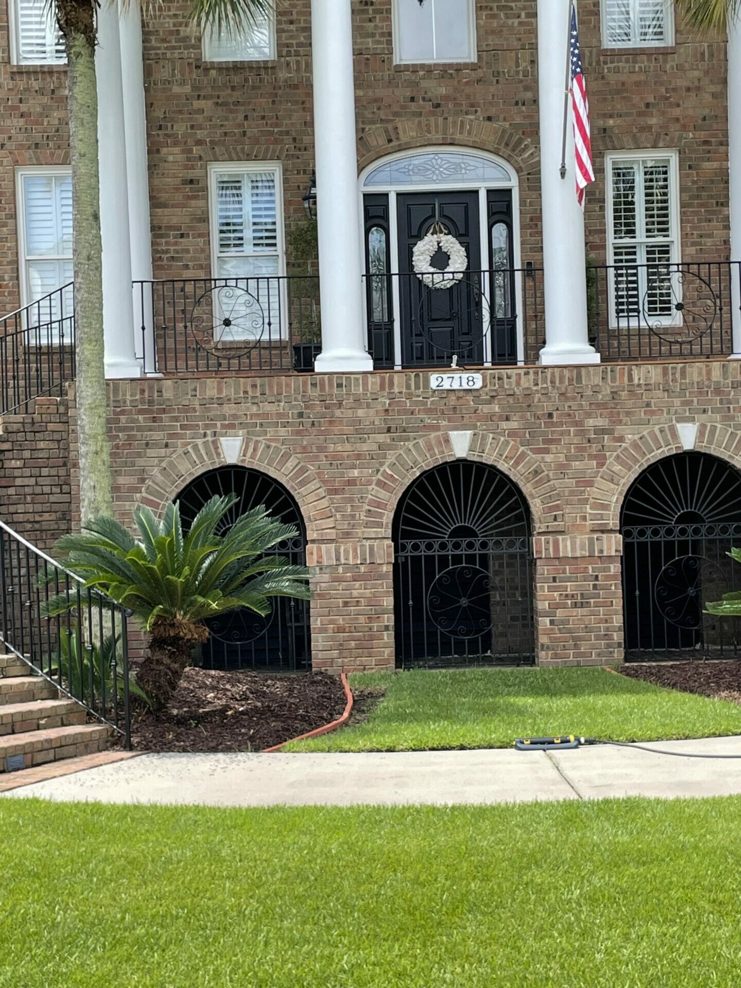 A brick house with white columns, a black front door with a wreath, the number 2718, an American flag, arched lower windows, and manicured green lawn with bushes features elegant wrought iron work on its balconies and iron decks.