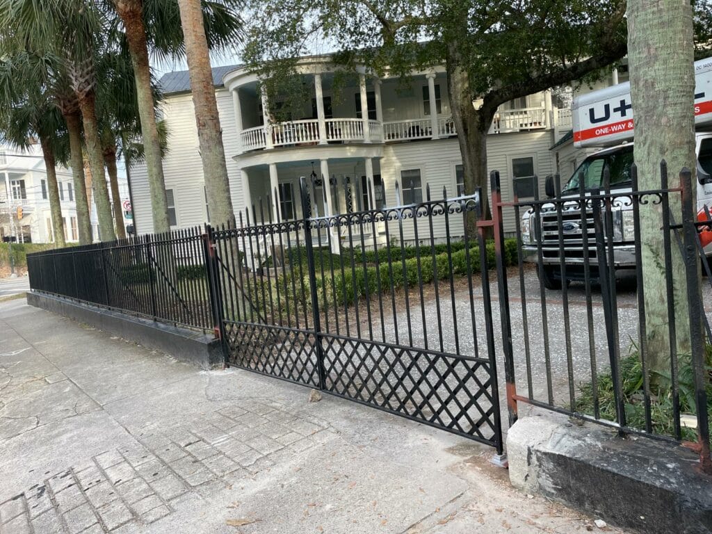 A black metal fence, showcasing classic Charleston iron work, is bent and damaged near the sidewalk in front of a large white house with columns and a U-Haul truck parked in the driveway.
