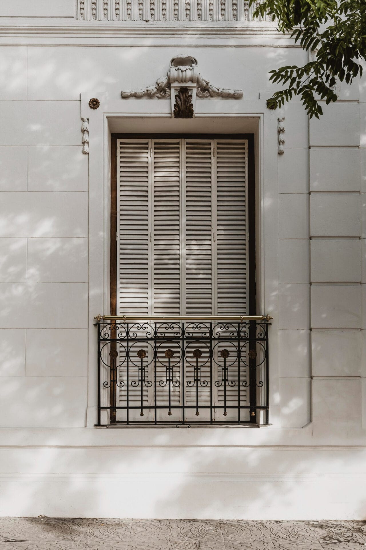 Closed white shutters on a window with an ornate black iron balcony railing, typical of Juliet balconies, on a white building facade.