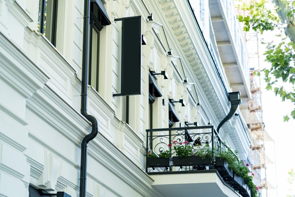 Ornate white building facade with black iron balcony, flower boxes, Juliet balconies, and blank vertical sign.