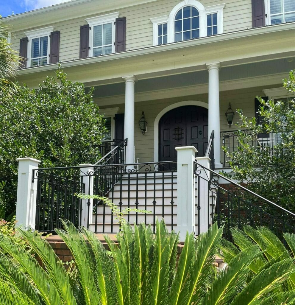 A two-story house with white siding, tall columns, and a staircase enhanced by elegant ironwork, surrounded by lush green plants.